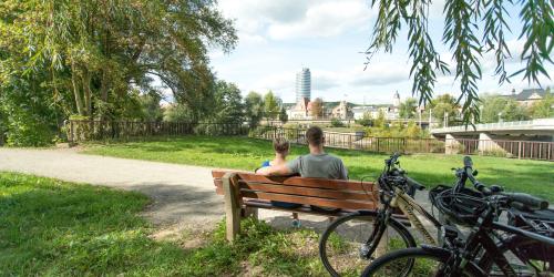 Im Fordergrund eine Bank mit 2 sitzenden Personen und angelehnten Fahrrädern, in der grünen Parklandschaft Oberaue mit Blick auf die Saale und die Stadtsilhouette  von Jena
