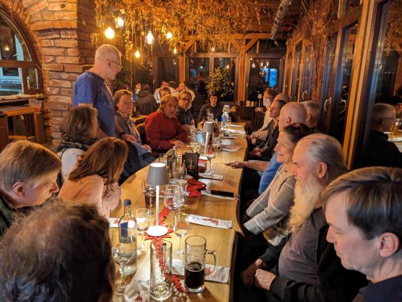 Viele Menschen sitzen in einem Raum an einem langen Tisch, gedämftes Licht, Jemand steht und spricht zu den anderen
