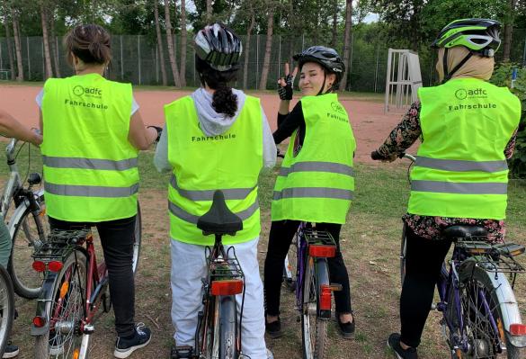 4 Frauen mit Warnwesten neongrün auf dem Fahrrad stehend von hinten fotografiert, eine Frau schut zum Fotografierenden