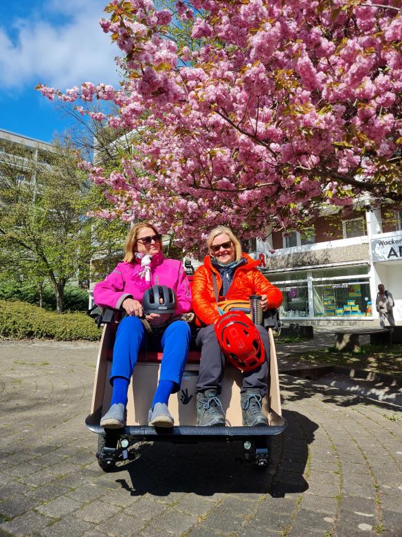 Im Vordergrund sitzen zwei farbenfroh gekleidete ältere Damen in einer Fahrradrikscha. Sie haben beide einen Fahrradhelm auf dem Schoß. Im Hintergrund Häuser und ein großer rosa blühender Baum.