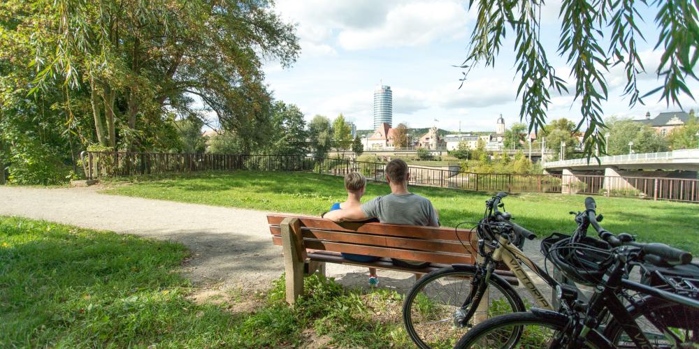 Im Fordergrund eine Bank mit 2 sitzenden Personen und angelehnten Fahrrädern, in der grünen Parklandschaft Oberaue mit Blick auf die Saale und die Stadtsilhouette  von Jena