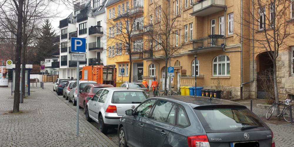 Stadtstraße mit parkenden Autos im Vordergrund, gepflasterter Gehweg, Bäume ohne Laub