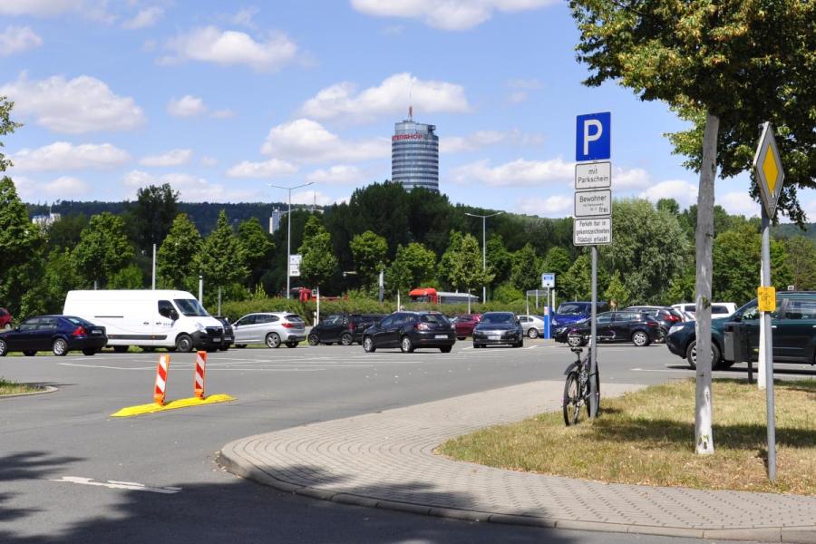 Blick auf den Seidelparkplatz in Jena im Vordergrunddie EInmündung, Bäume einzenlne Fahrzeuge, im Hintergrund ist der Uniturm zu sehen.