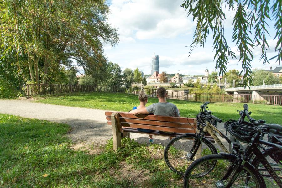Im Fordergrund eine Bank mit 2 sitzenden Personen und angelehnten Fahrrädern, in der grünen Parklandschaft Oberaue mit Blick auf die Saale und die Stadtsilhouette  von Jena