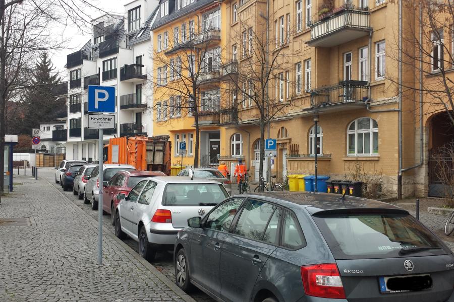 Stadtstraße mit parkenden Autos im Vordergrund, gepflasterter Gehweg, Bäume ohne Laub